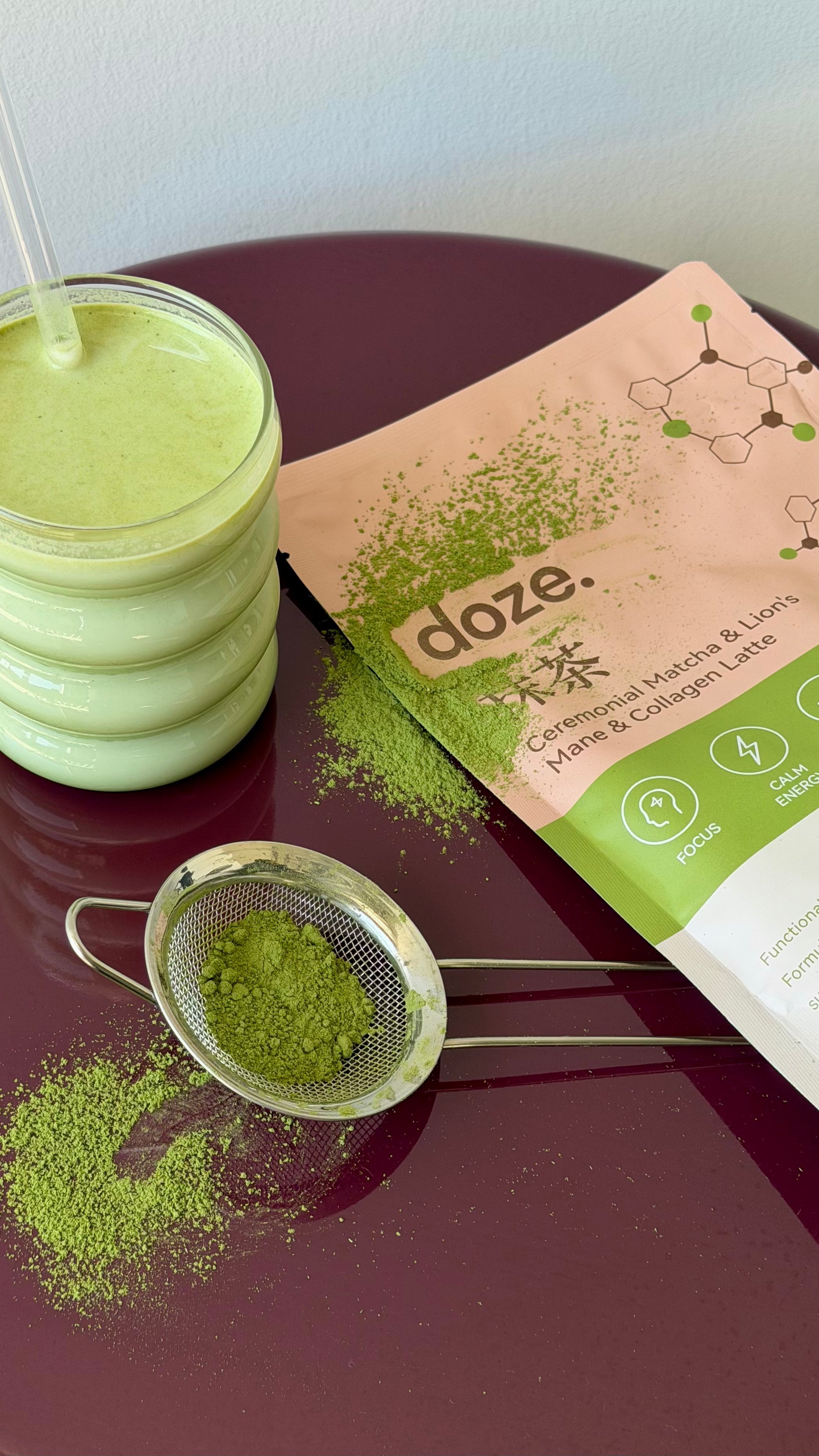 doze. matcha pouch on a burgundy table with a glass of matcha latte and a sieve of loose matcha powder.