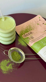 doze. matcha pouch on a burgundy table with a glass of matcha latte and a sieve of loose matcha powder.