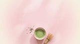 Bowl of whisked ceremonial matcha with a bamboo whisk and loose green tea leaves on a blush pink background