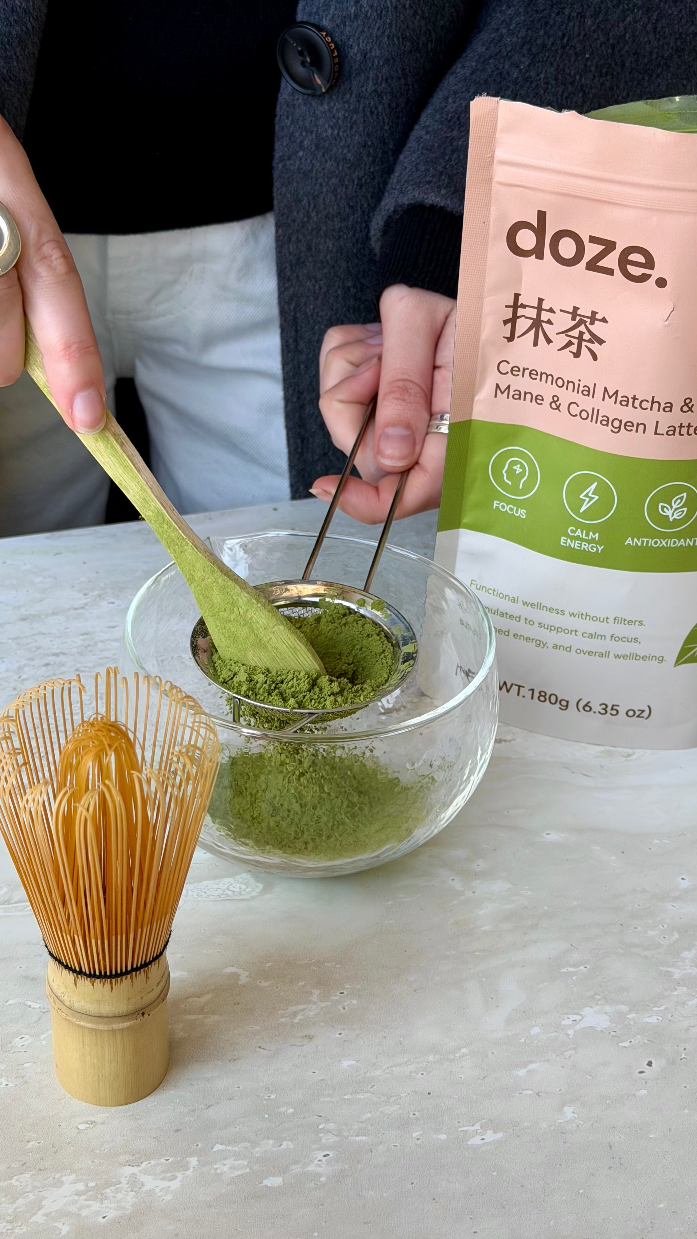 Hands sifting doze. matcha powder into a glass bowl beside a bamboo whisk and the doze. pouch.