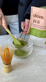 Hands sifting doze. matcha powder into a glass bowl beside a bamboo whisk and the doze. pouch.