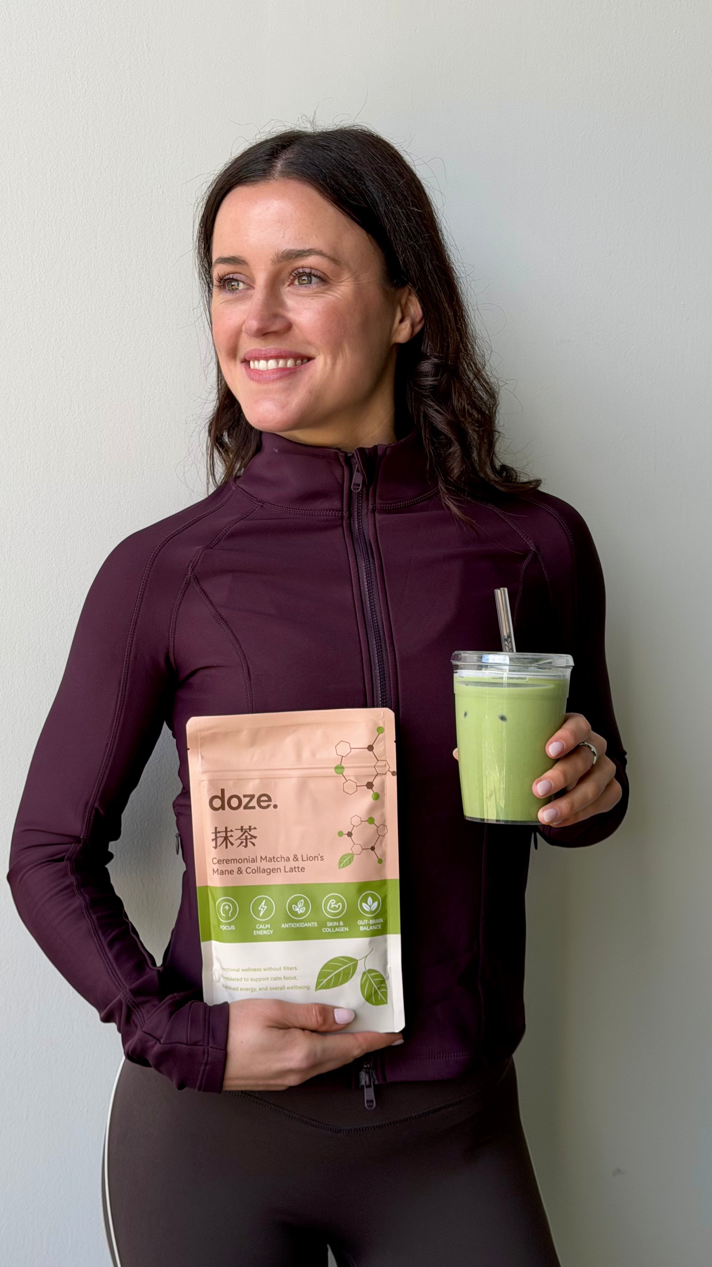 Woman in a plum zip-up jacket holding a doze. matcha pouch and a clear tumbler of iced matcha latte.