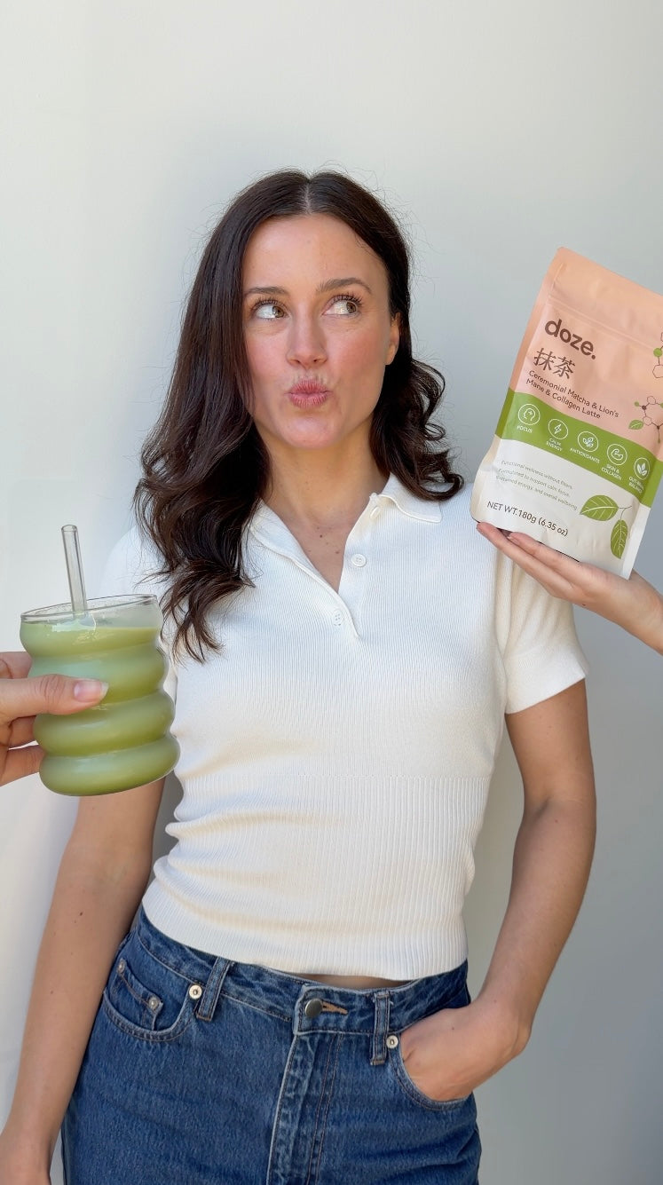 Woman in a white top holding up a doze. matcha pouch beside a glass of iced matcha latte.
